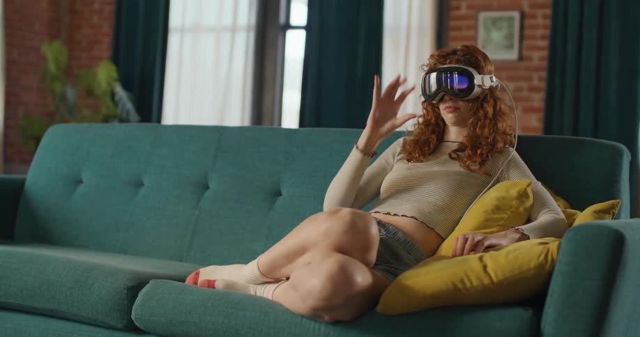 A young woman sitting on a couch wearing a virtual reality headset and using one hand to select something