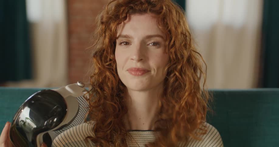 The video is a closeup shot of a young woman with curly red hair removing a white virtual reality vr headset with dark lenses from her face after taking off the device she smiles softly at the camera and touches her hair