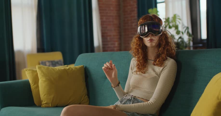 Teenage girl sitting on the couch wearing a virtual reality headset as she moves her arms and looks around