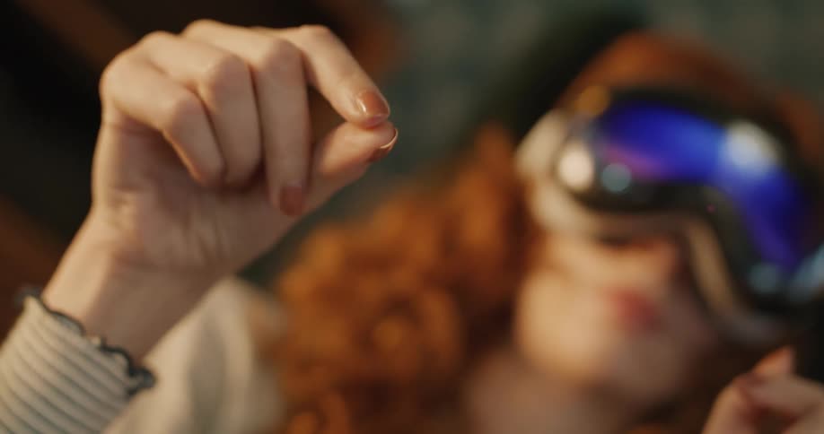 A close view of a red haired woman wearing a virtual reality headset shows her hand interacting with a virtual object by pinching her fingers in the foreground