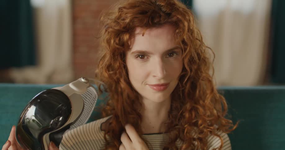 A woman with curly red hair is initially seen wearing a virtual reality vr headset with dark blue lensesshe removes the headset smiles adjusts her hair and continues to hold the device in her hand
