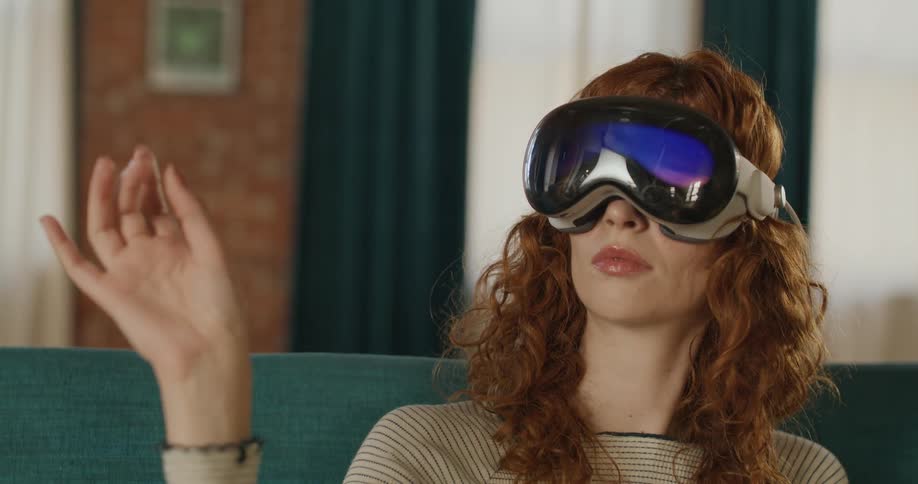 A young woman with long brown hair sits on a couch at midday wearing a vr headset and using hand gestures to operate it