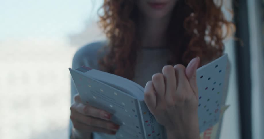 A woman with curly red hair sitting by a bright window opens and reads a book titledthe secret lives of colorwith a polka dot cover