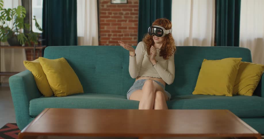 Teenage girl sitting on the couch wearing a virtual reality headset as she moves her arms and looks around