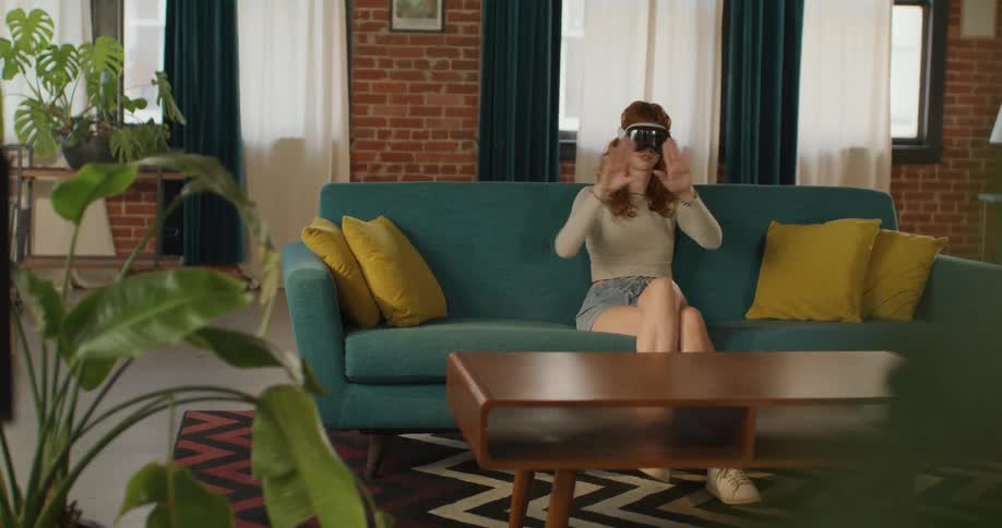 A woman with curly red hair sits on a teal sofa in a loftstyle room with exposed brick wears a dark visor vr headset and moves both hands and arms in front of her face interacting with the technology interface