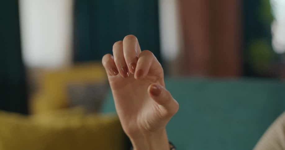 A close view shows a womans hand with light brown nail polish making a subtle pinching or tapping gesture in a brightly lit room with blurred blue and yellow furniture in the background