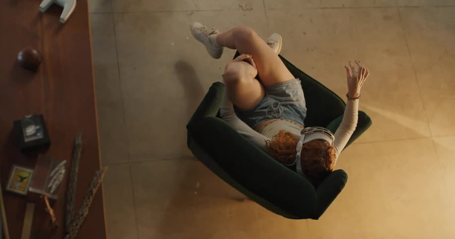 A woman with red hair lies backward in a dark armchair with legs bent and raised moves her arms above her head and uses a vr headset the ambiance has a tiled floor near a wooden surface with small objects