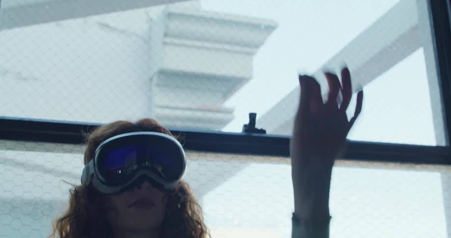A young woman wearing an apple vr headset stands beside a window at midday using her finger to make gestures