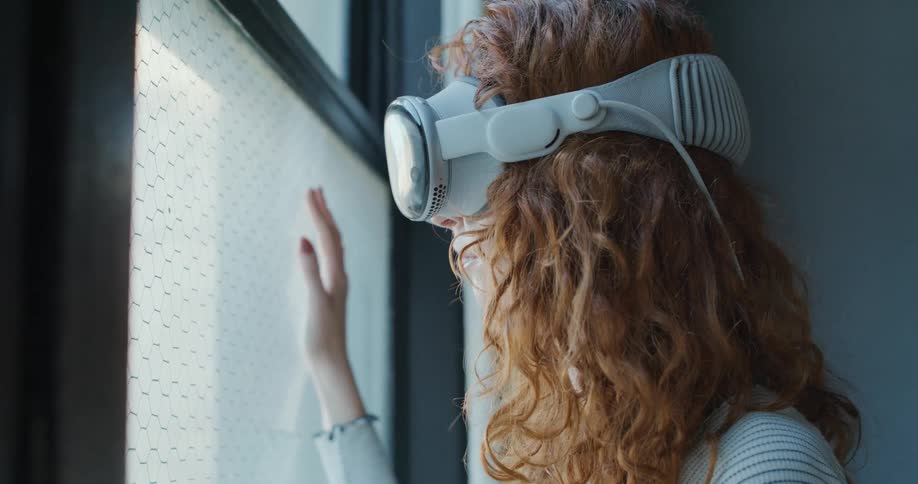 The video is a medium closeup shot of a woman with curly red hair wearing a white vrmr headset looking out a window she is seen raising her right hand and resting it lightly on the glass of the window as if touching a virtual display or projection