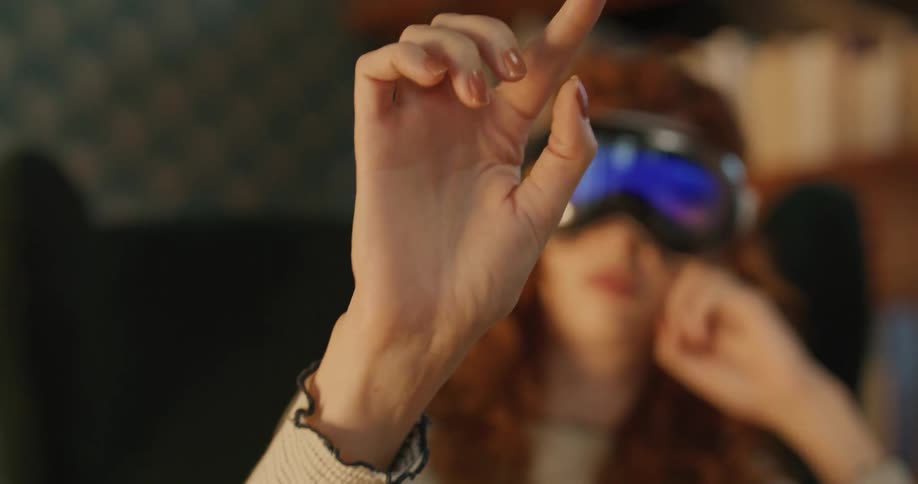 A woman with curly red hair sits in a chair wearing a vr headset uses one hand to make tapping and pinching motions in the air and rests her other hand near her ear in a dimly lit room with bookshelves