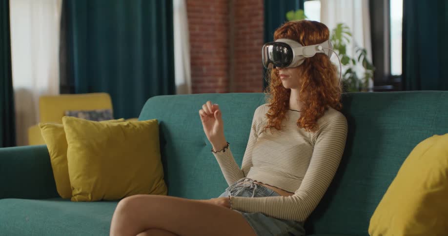 A young woman sitting on a couch wearing a virtual reality headset and moving her hand to select something