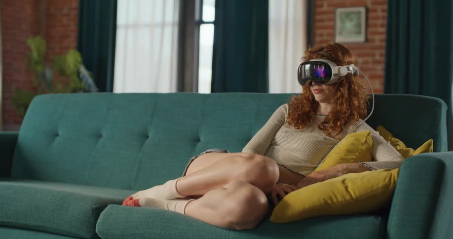 A red haired woman wearing a white long sleeve shirt shorts and a virtual reality headset reclines on a teal sofa with yellow pillows in a bright living room