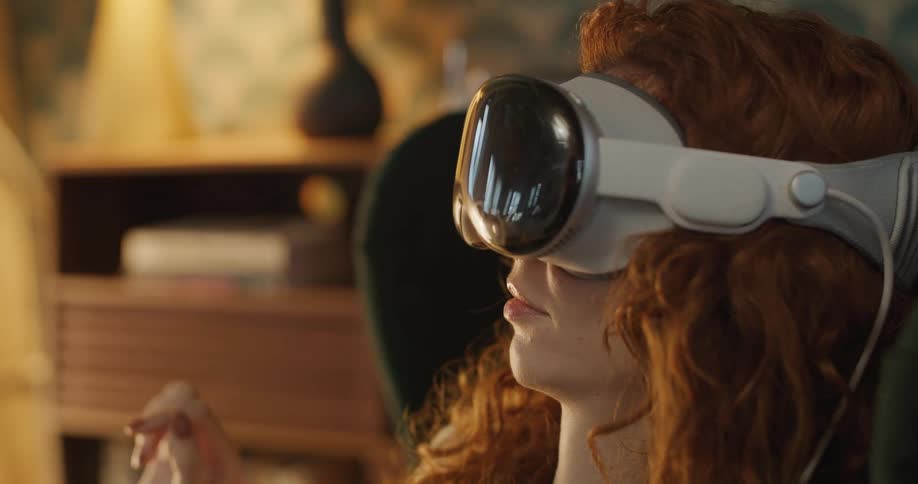 A woman with wavy red hair wears a virtual reality headset her hand raised in a gesture of interaction a slightly blurred furniture visible in the background