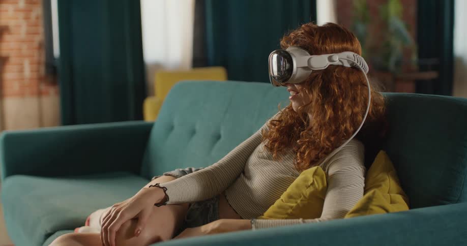 A woman with long curly red hair relaxes on a turquoise couch wearing a light colored long sleeve shirt and a white virtual reality headset in a brightly lit living room