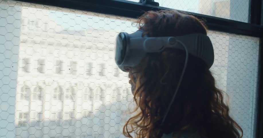 A person with long dark hair is shown from behind initially wearing a sleep mask while looking out a window at a city buildingthe person then lowers the sleep mask and replaces it with a virtual reality vr headset continuing to look out th