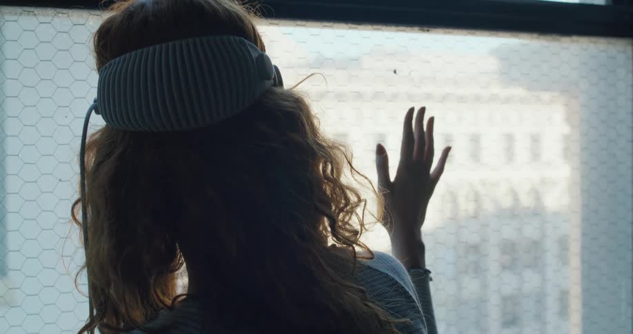 A woman with curly hair wearing a ribbed vr headband stands at a wiremeshed window with her hand raised toward the glass