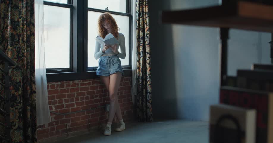 A female teenager reading a book while standing by the window in a room during daytime