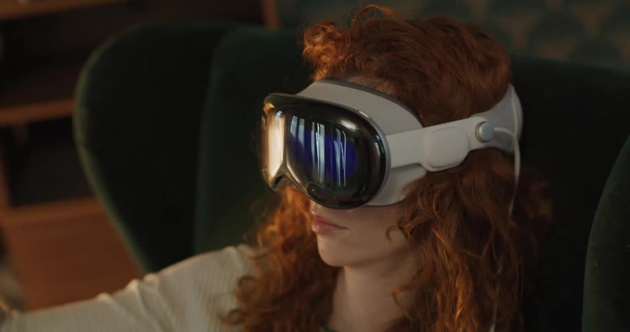 A close side view shows a red haired woman wearing a white shirt and a sleek virtual reality headset reaching out her hand to interact with a virtual environment in a dimly lit room