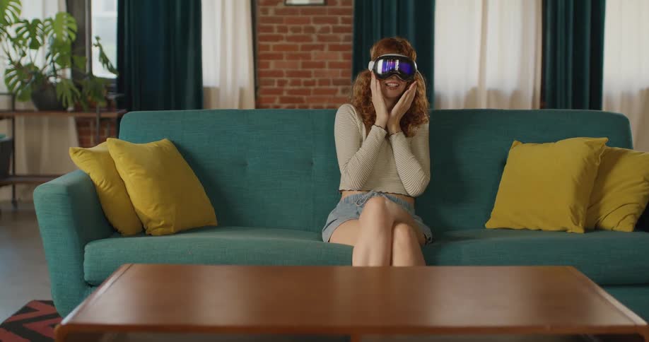 A young woman sitting on a couch in the living room wearing a virtual reality headset moving her hands and getting startled by something she saw