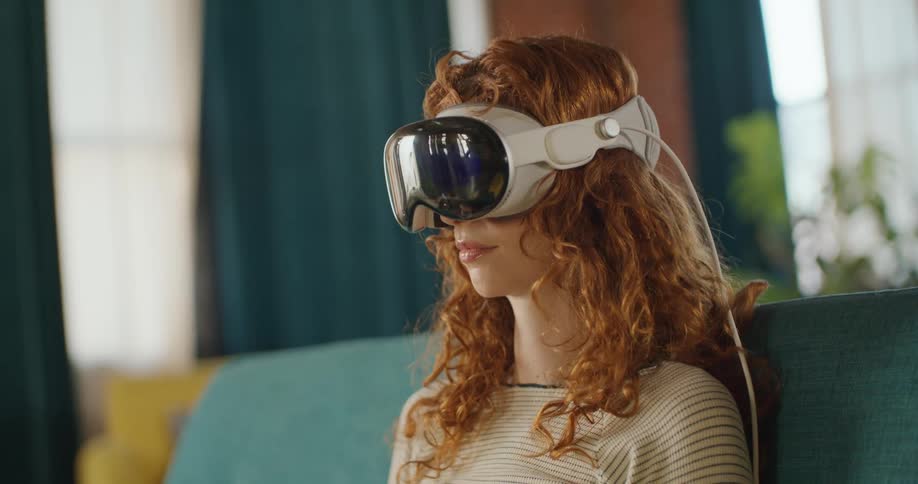 A young woman sitting on a couch in the living room wearing a virtual reality headset moving her hands and getting startled by something she saw