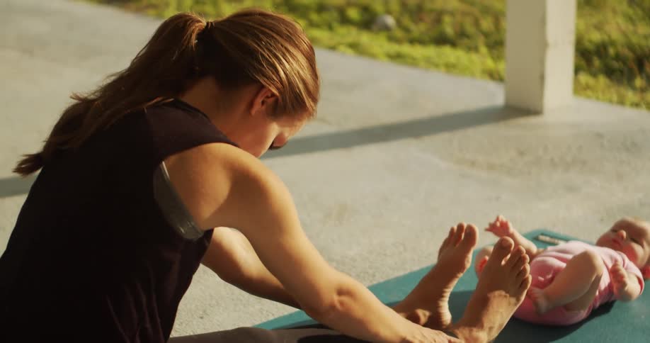 A woman stretches on a yoga mat where her baby lays on its back