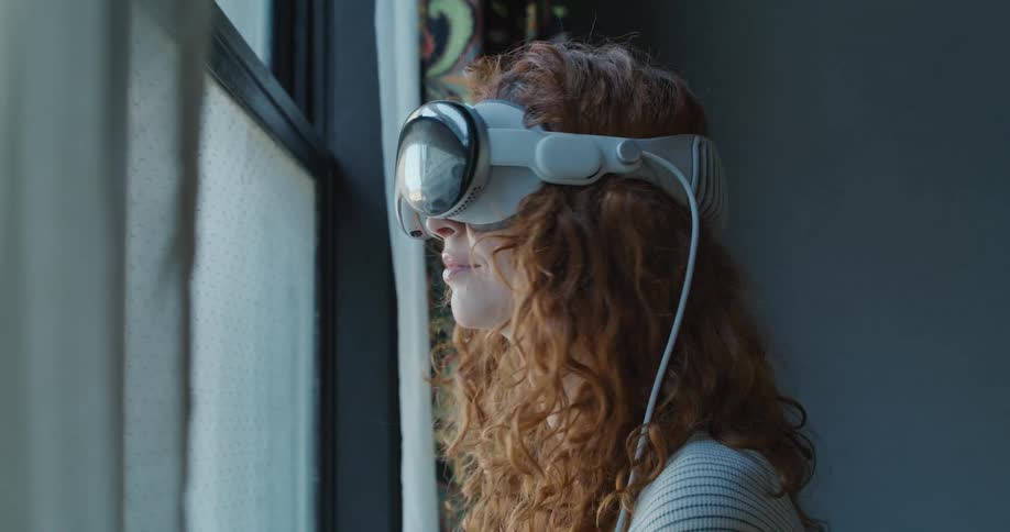 A woman with curly red hair wears a white vr headset stands near a window with sheer curtains and looks forward and upward engaging with a perceived environment while smiling