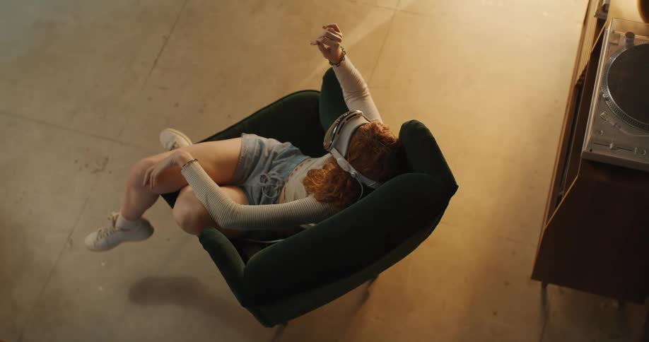 An overhead view shows a red haired woman wearing a virtual reality headset a long sleeve shirt and shorts leaning back in a dark green armchair with her arms and legs outstretched as if dancing or reacting to the virtual world