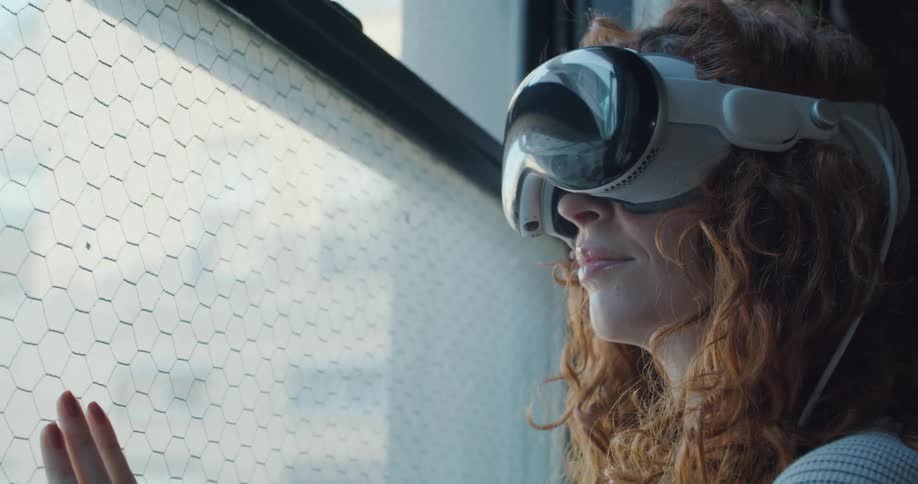 A woman with curly red hair wears a futuristic white vr headset and looks out a window with a faint smile her hand resting on the wire mesh glass