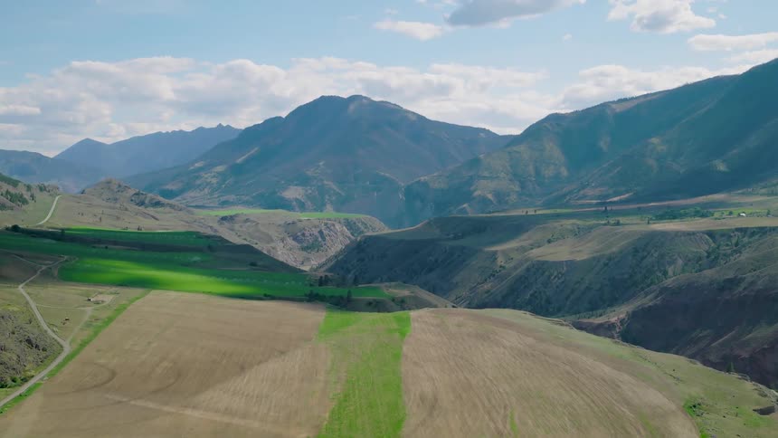 A highangle aerial view captures a vast mountain valley and its deep gorge with fluffy clouds drifting slowly over the distant peaks and patchwork fields
