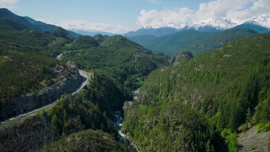 An aerial wide shot shows a winding highway and a river running through a steep green wooded canyon with sharp mountain peaks capped with white snow in the distance