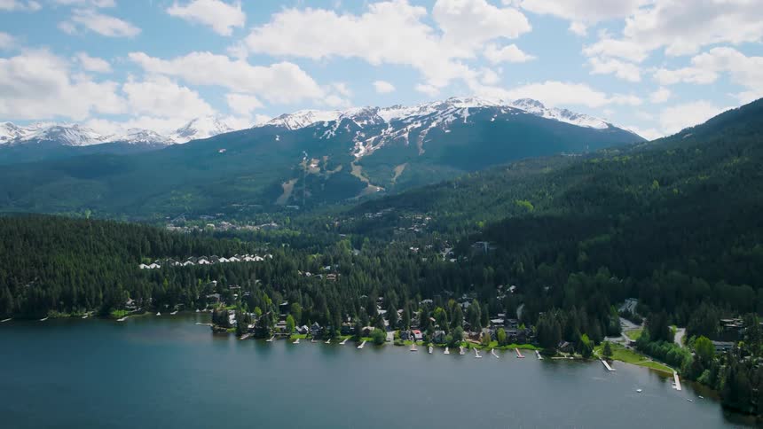 An aerial view shows a dark blue lake bordered by a shoreline of green pine trees and houses with large snow capped mountains under a bright cloudy sky