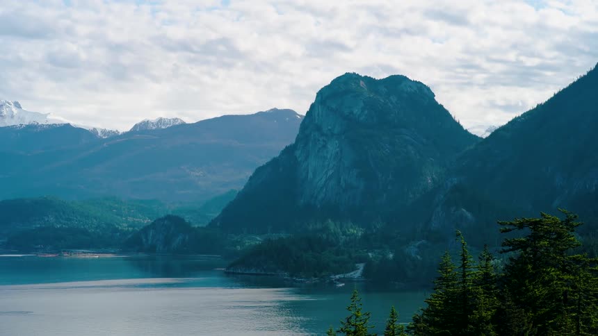 A stunning mountain range rises over a calm blue lake as soft white clouds drift quickly across the sky in a timelapse