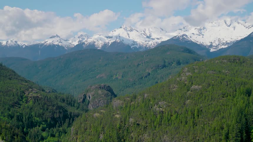 Aerial view moving over forested mountains with snow covered mountains in the distance