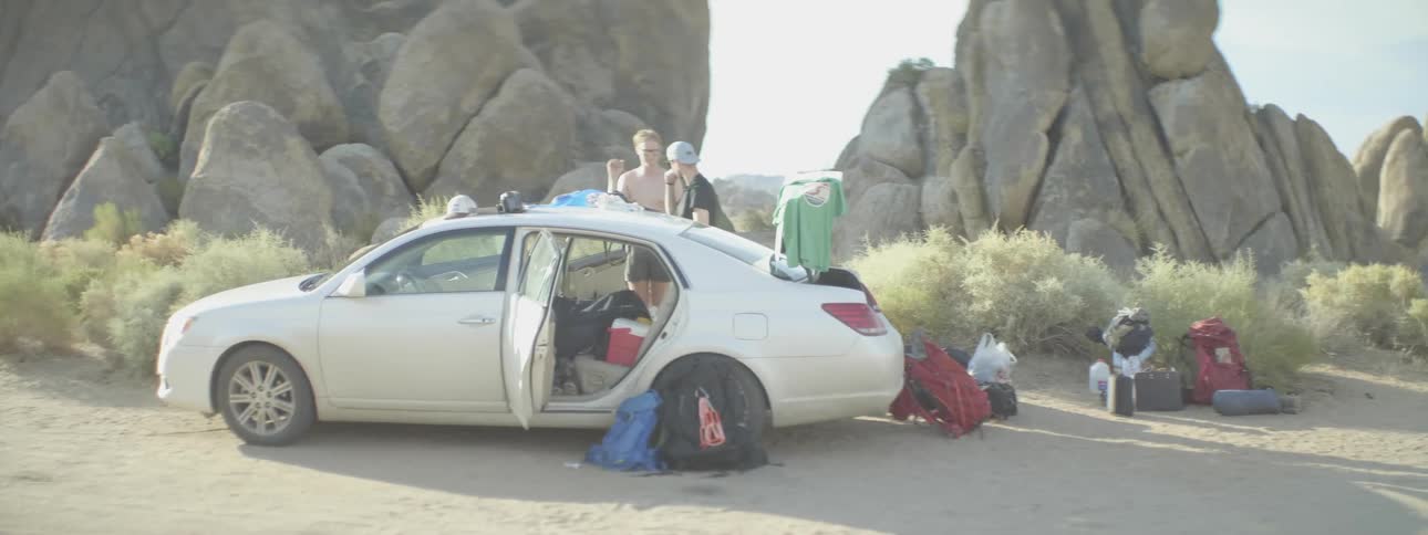 Two young men stand outside their vehicle with all their camping gear
