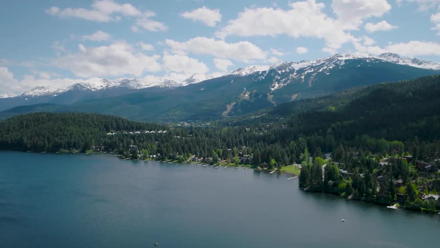 A wide body of water sits in a valley bordered by dense conifer forests containing small residential structures and tall snowcovered mountains rise steeply in the background under a blue sky with scattered clouds