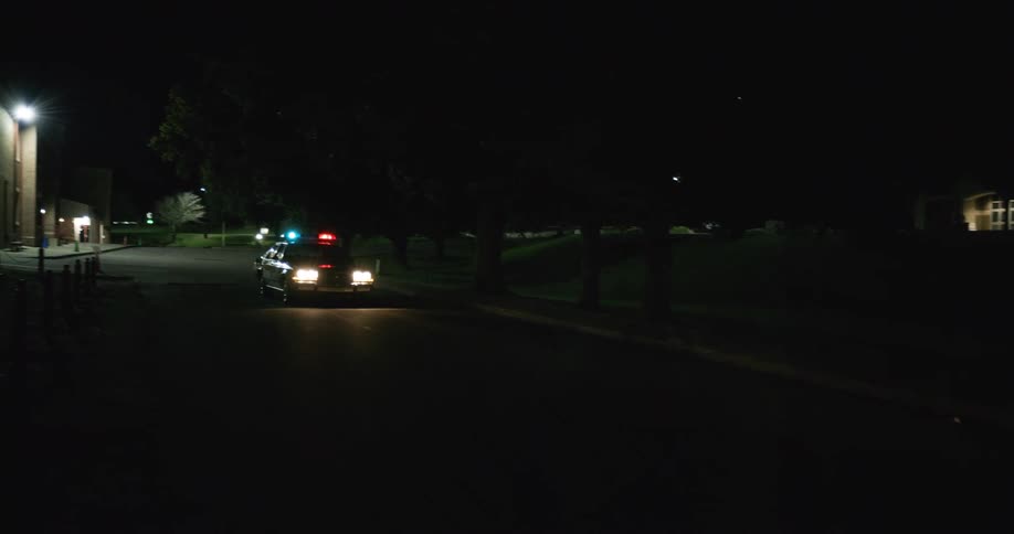 The video shows a police car with its blue and red lights flashing driving down a dark road at nightthe vehicle passes a large brick building on the left and continues down the suburban street before speeding past the camera