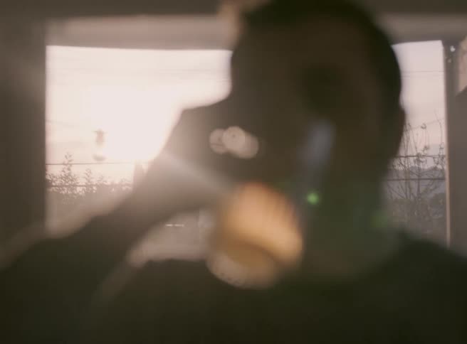 A person stands indoors with his head and shoulders silhouetted in the foreground while drinking beverage in a glass facing outward through a window toward a bright hazy hillside urban landscape crisscrossed with electrical wires and poles