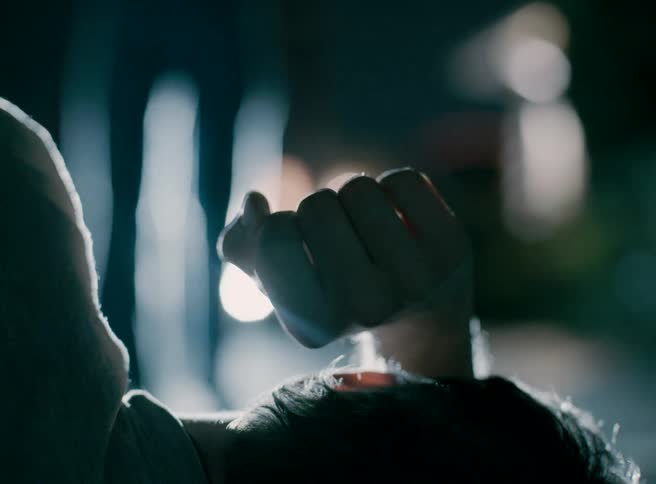 This is a dramatic lowangle closeup of a persons clenched fist and head in dark silhouette against a blurry night background bright outoffocus headlights or streetlights flash and flare behind them creating a sense of urgency or distress