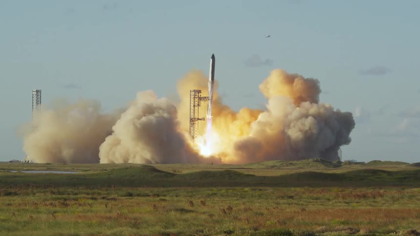 A space rocket lifts off from an open field into the sky at midday