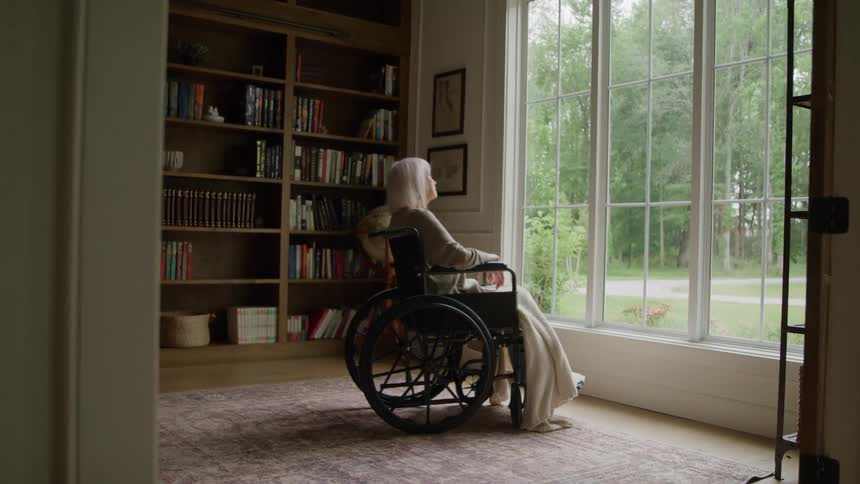 Female in a wheelchair looking out to yard from large picture window in the house