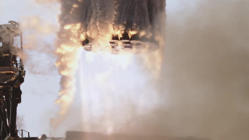 The video is a dramatic closeup shot of a large rocket engine during launch with multiple nozzles firing and producing a dense cascade of fiery exhaust and white smoke the plumes of flame and smoke are seen moving in a powerful turbulent rus