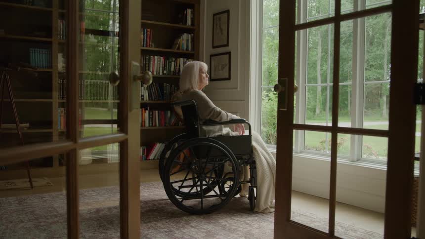 The video shows a senior woman in a wheelchair sitting indoors looking out a large window into a lush green yardthe room features a wall lined with bookshelves and french doors in the foreground suggesting a comfortable library or sitting roo