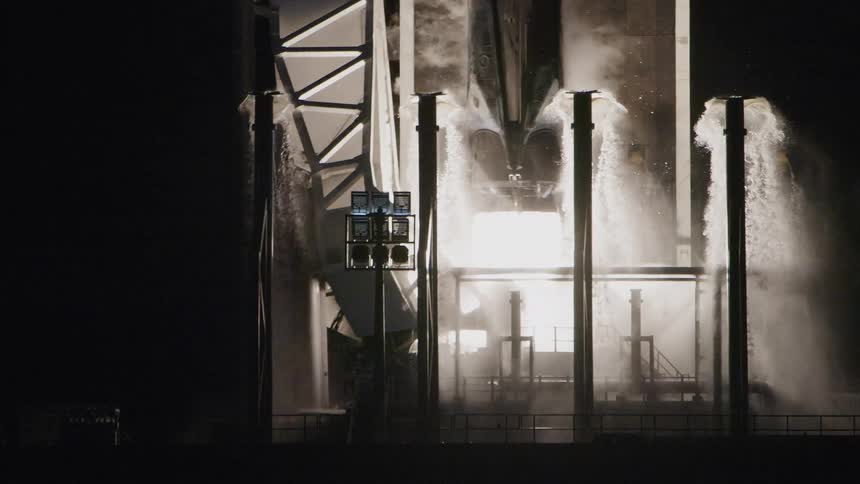 The base of a rocket stands on a launch pad a water suppression system sprays streams of water from vertical pillars and the main engine ignites producing a bright plume of exhaust and smoke
