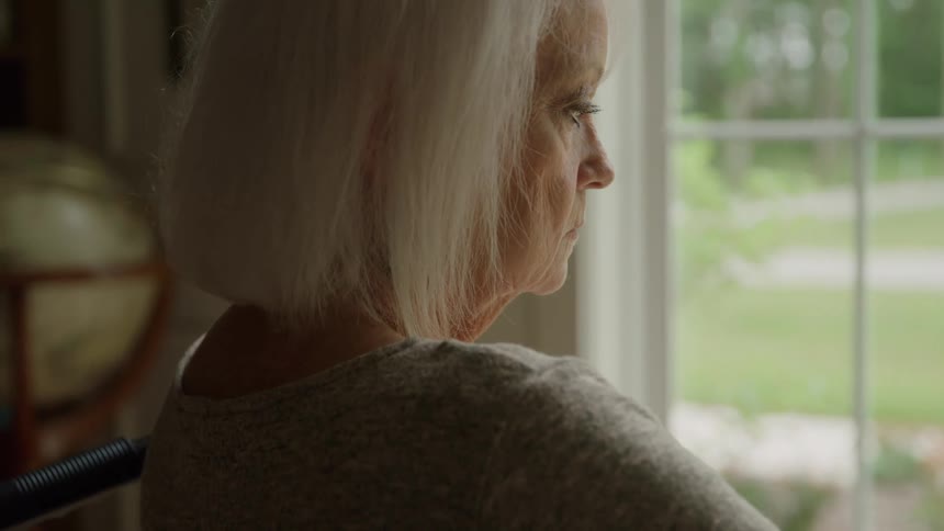A close side view shows an elderly woman with white hair looking out a large window at the bright green outdoors from inside a dimly lit room