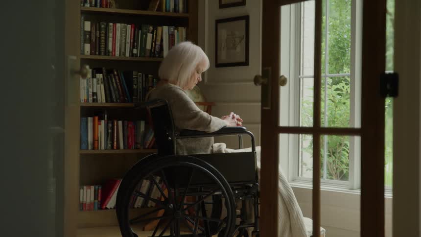 An elderly woman sits in a wheelchair by a window in a home library