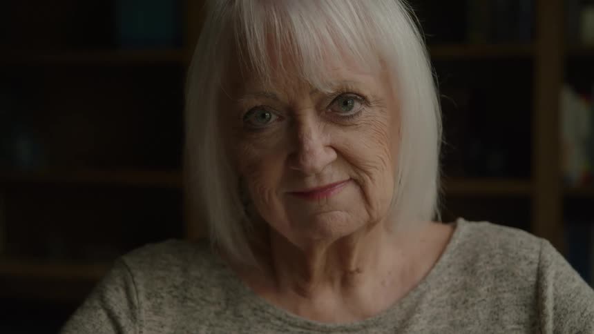A close dimly lit view shows a serene older woman with bright white hair and bangs wearing a gray shirt looking directly at the camera with a gentle smile against a dark blurred bookshelf background
