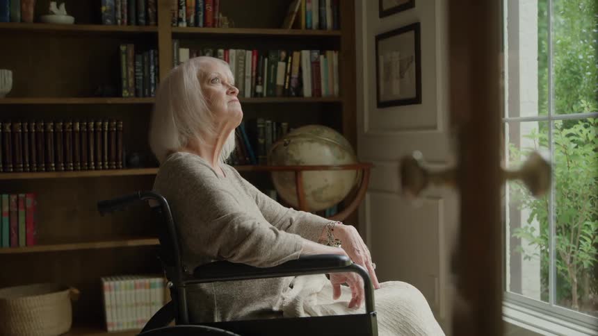An elderly woman sitting on a chair in front of a window at her home silently observing