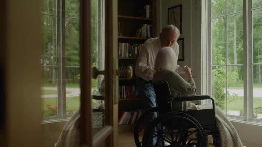 An elderly woman in a wheelchair looking out the window as an old man arrives with coffee cups hands her one and then kisses her forehead