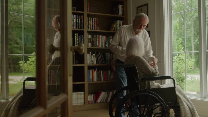 An elderly man brings a cup to his wife who is sitting in a wheelchair and looking out a large window and they both drink together in a quiet book lined room