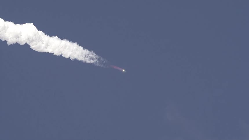 A small rocket with a visible engine glow flies quickly across the center of a deep blue sky leaving a long dense white cloud of exhaust that dissipates slowly